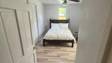 Un couloir étroit mène à une chambre à coucher qui contient un lit, un ventilateur de plafond et une fenêtre.