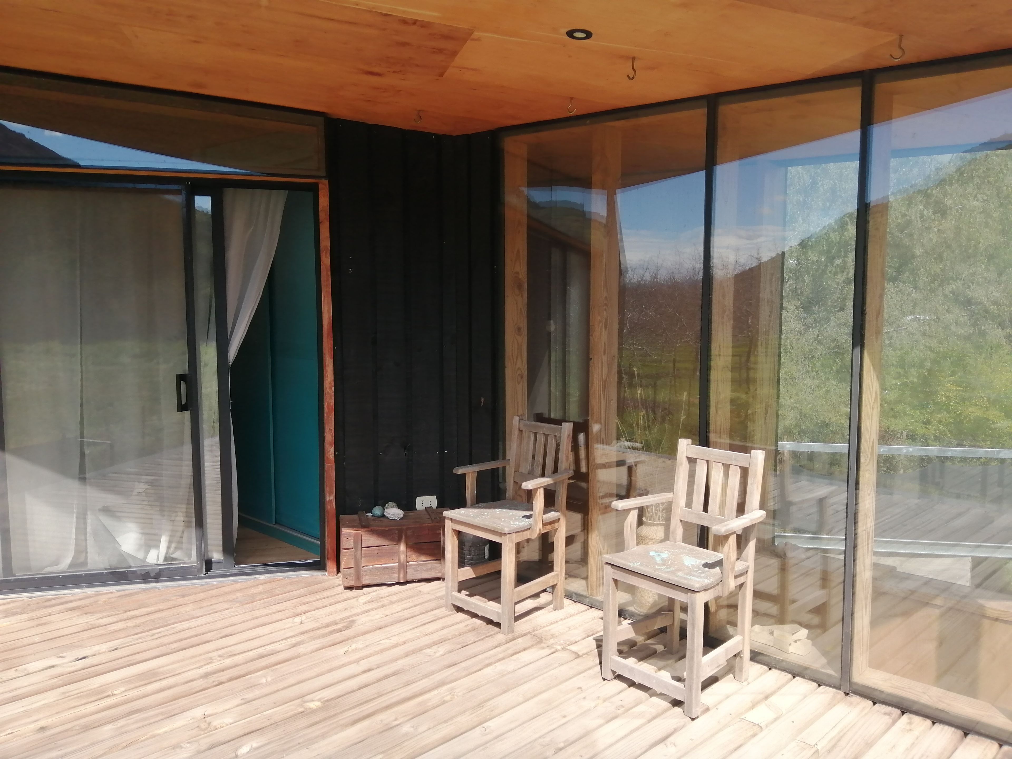 A wooden deck with chairs and a table, large glass doors, and a view of greenery.