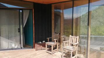 A wooden deck with chairs and a table, large glass doors, and a view of greenery.