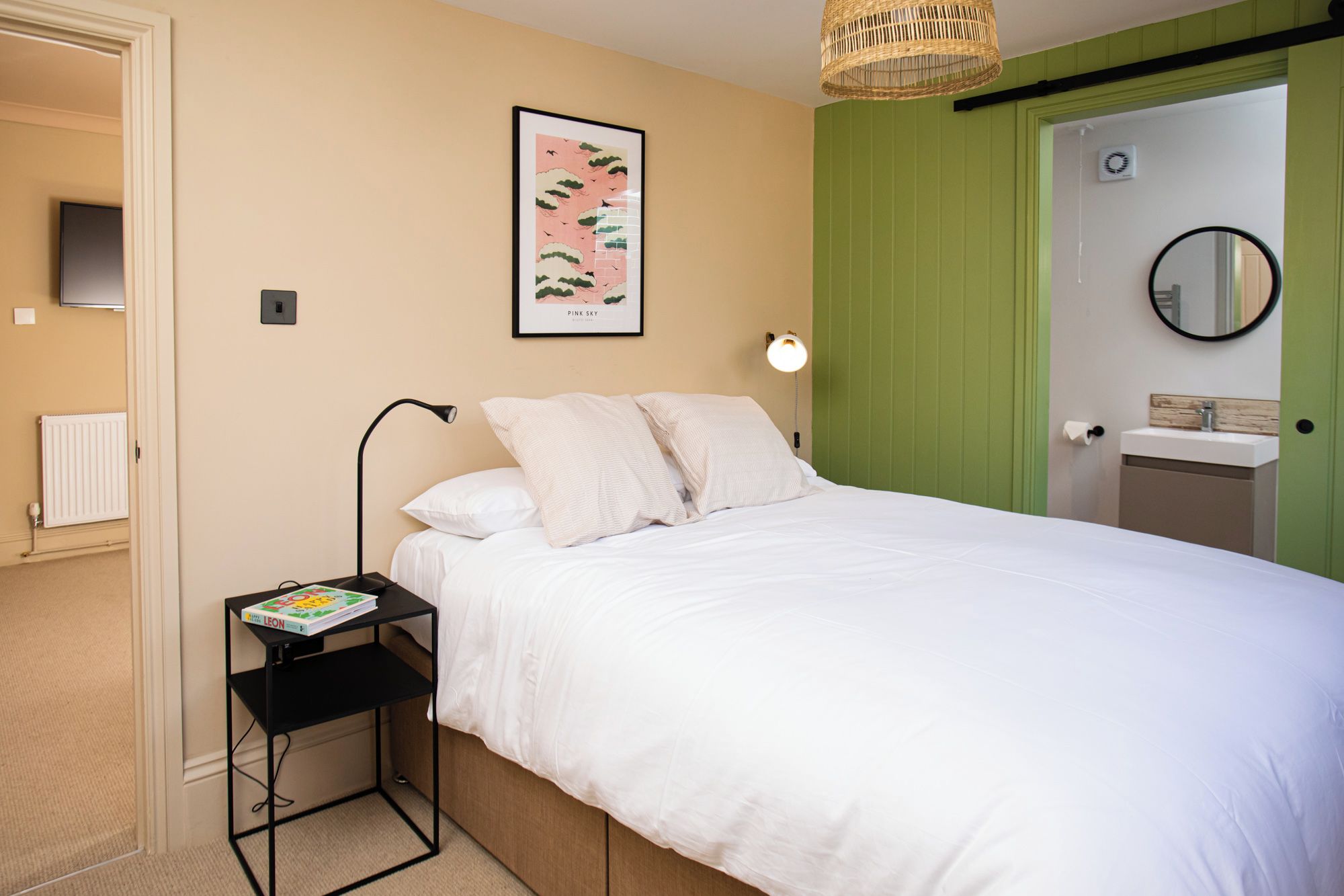 Une chambre d’hôtel avec un lit, une table de chevet sur laquelle se trouve un livre, un tableau accroché au mur, une lampe de chevet, une porte verte, un miroir et un radiateur blanc.