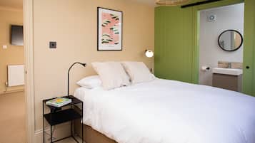 A hotel room with a bed, a bedside table with a book, a wall-mounted picture, a bedside lamp, a green door, a mirror, and a white radiator.