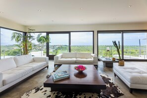 Ein modernes Wohnzimmer mit einem weißen Sofa, einem Couchtisch und Blick auf eine grüne Landschaft durch große Fenster.