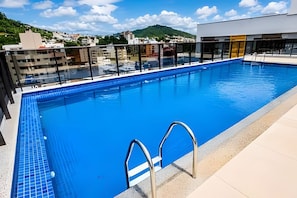 A swimming pool with clear blue water, surrounded by a modern building with glass railings. - Studio em ultimo andar linda vista (Florianópolis)