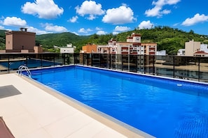 A rooftop swimming pool with clear blue water, surrounded by buildings and a mountain in the background.