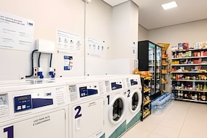 A laundromat with multiple washing machines, a self-service checkout, and a variety of packaged goods on shelves. - Studio em ultimo andar linda vista (Florianópolis)
