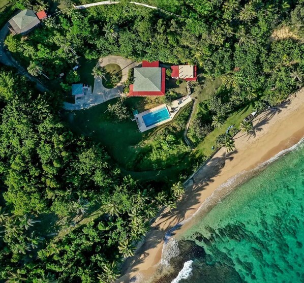 Vue aérienne d’une propriété située sur la côte, avec une piscine, un bâtiment à toit rouge et une plage.