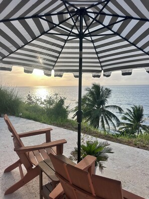 A beachside seating area with a striped umbrella, wooden chairs, and a view of the ocean at sunset.