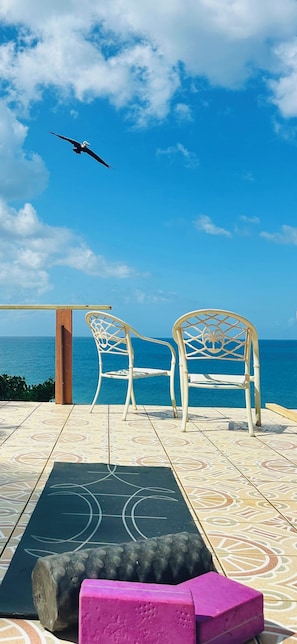 Une terrasse avec deux chaises blanches, un tapis noir et un bloc rose, d’où l’on peut admirer un ciel bleu azur et l’océan.