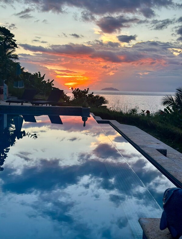 A pool with a view of the sunset over the ocean, surrounded by greenery and lounge chairs.