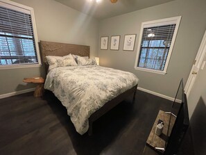 A bedroom with a bed, a nightstand, a wooden stool, framed botanical prints on the wall, and a television on a stand. - Fishing Frenzy Funhouse: Hot Tub/Lakefront Fishing/15 Minutes to Attractions! (Sevierville)
