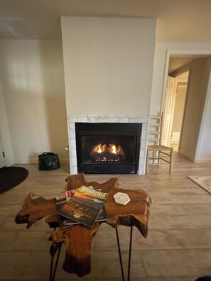Een kamer met een open haard, een leren poef met boeken, en een houten vloer.