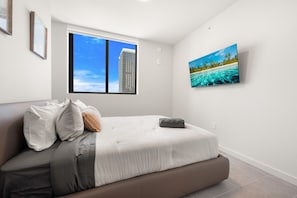 Un dormitorio con cama, almohadas, una ventana con vistas a la ciudad y un cuadro colgado en la pared que representa una playa.