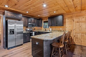 A modern kitchen with a stainless steel refrigerator, a microwave, and a granite countertop island with bar stools. - Riverfront A-Frame | Hot Tub + Fireplace (Ellijay)