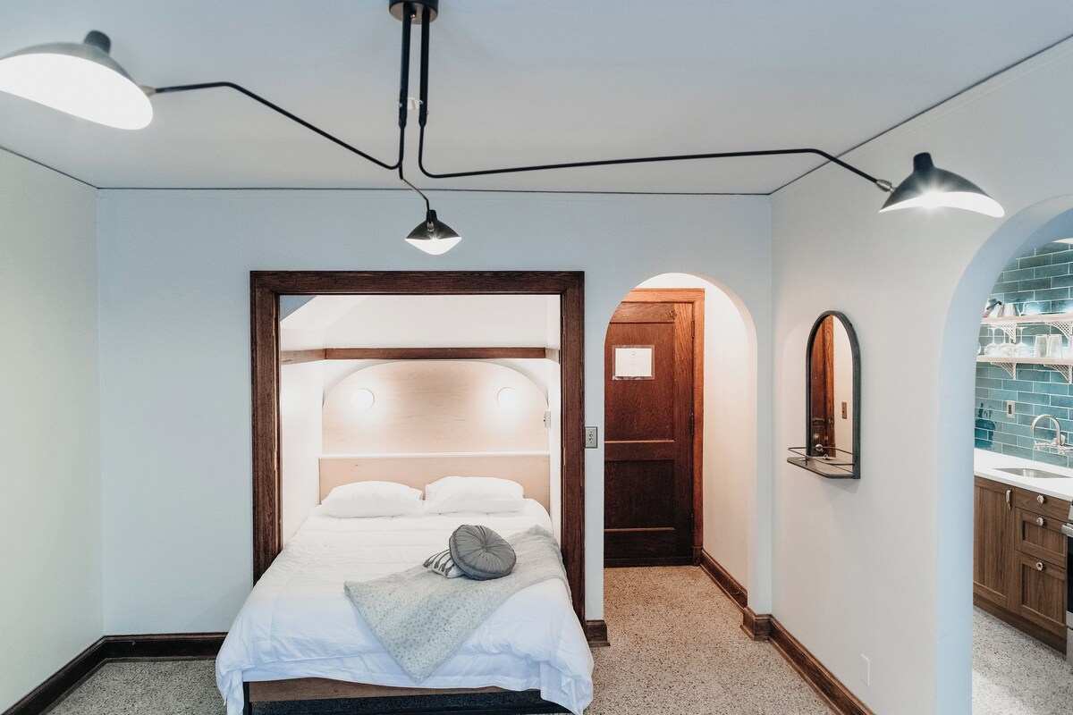 A bedroom with a bed, a bedside table, a mirror, and a view of a bathroom.