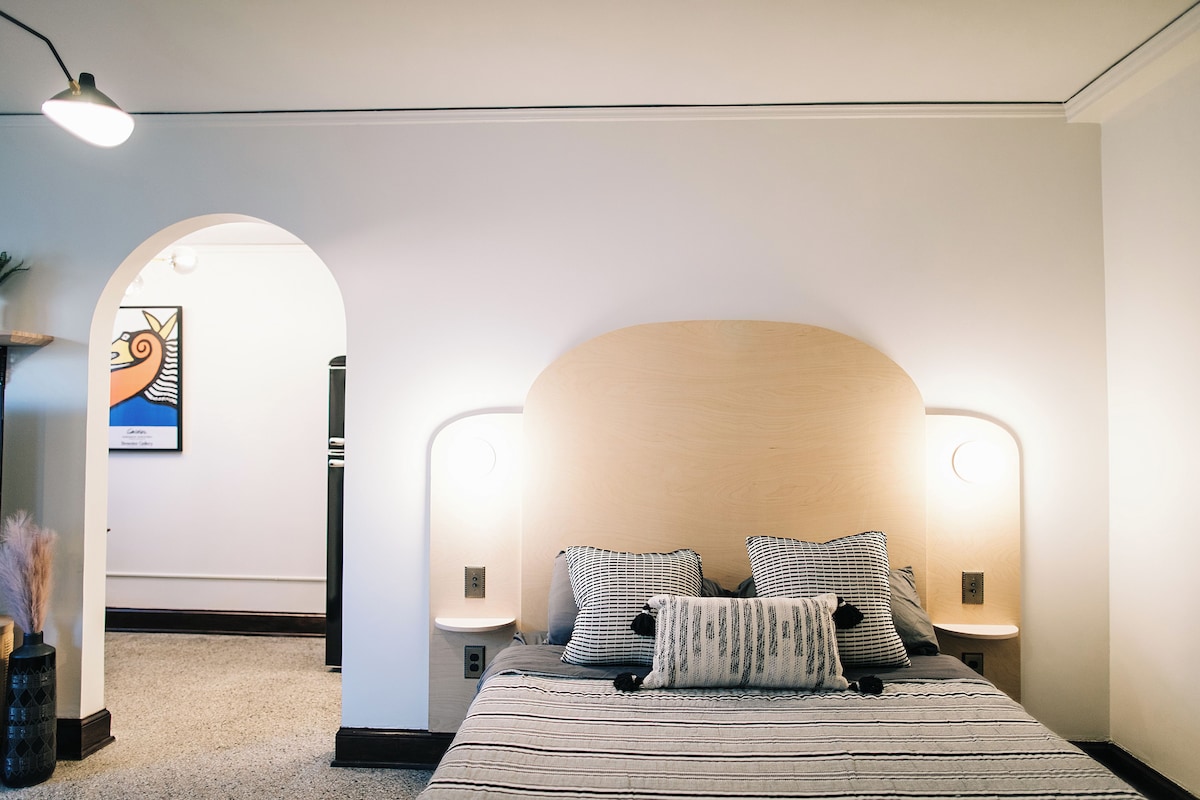 A bedroom with a large bed, two bedside lamps, a wall-mounted light, and a framed artwork on the wall.