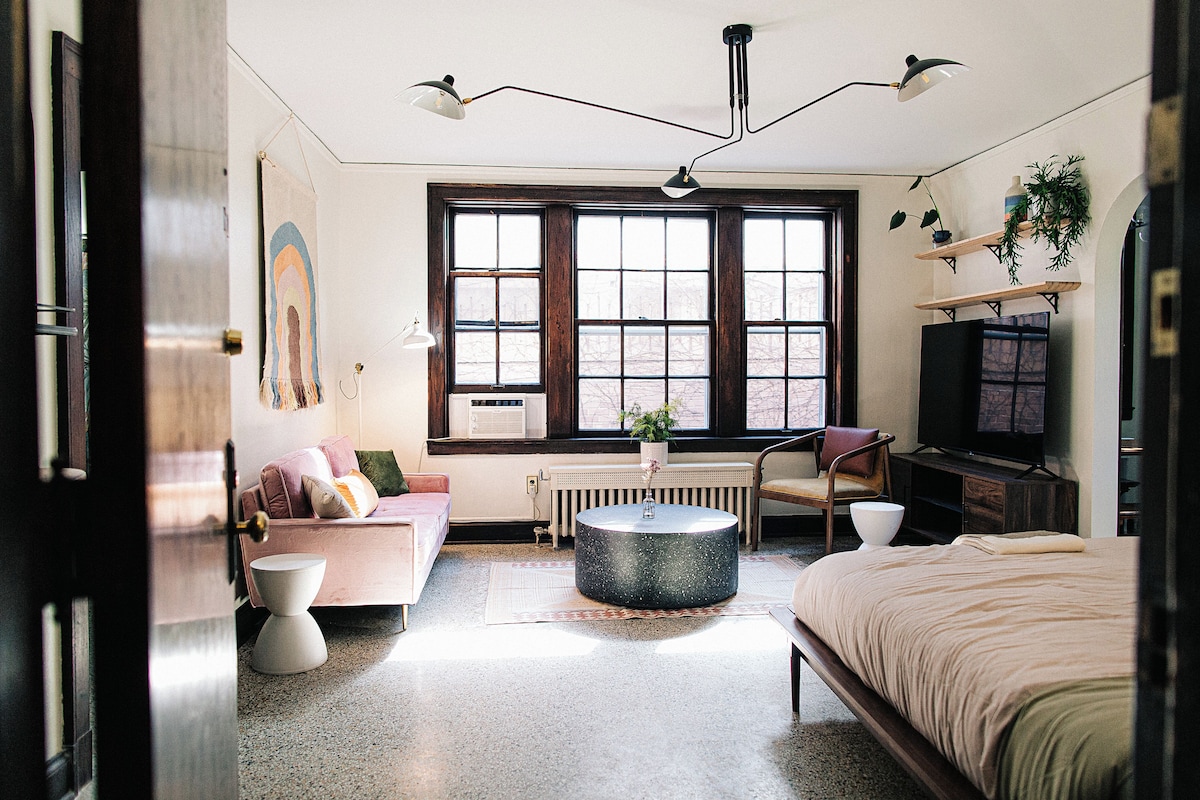 A room with a bed, a sofa, a coffee table, a television, and a window with a view of the outside.