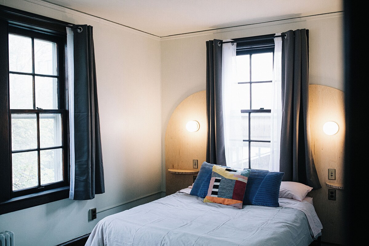 A hotel room with a bed, two windows with curtains, and wall-mounted lights.