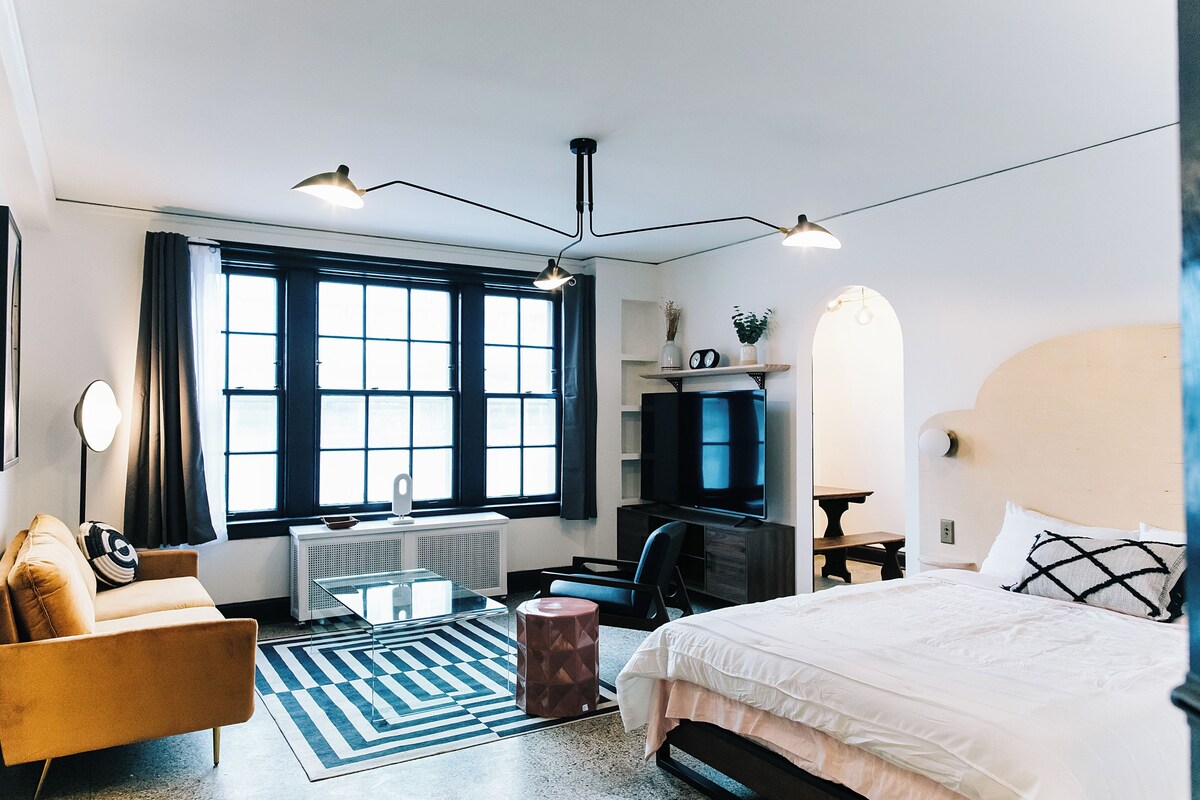 A modern hotel room with a bed, a desk, a chair, a TV, and a large window.