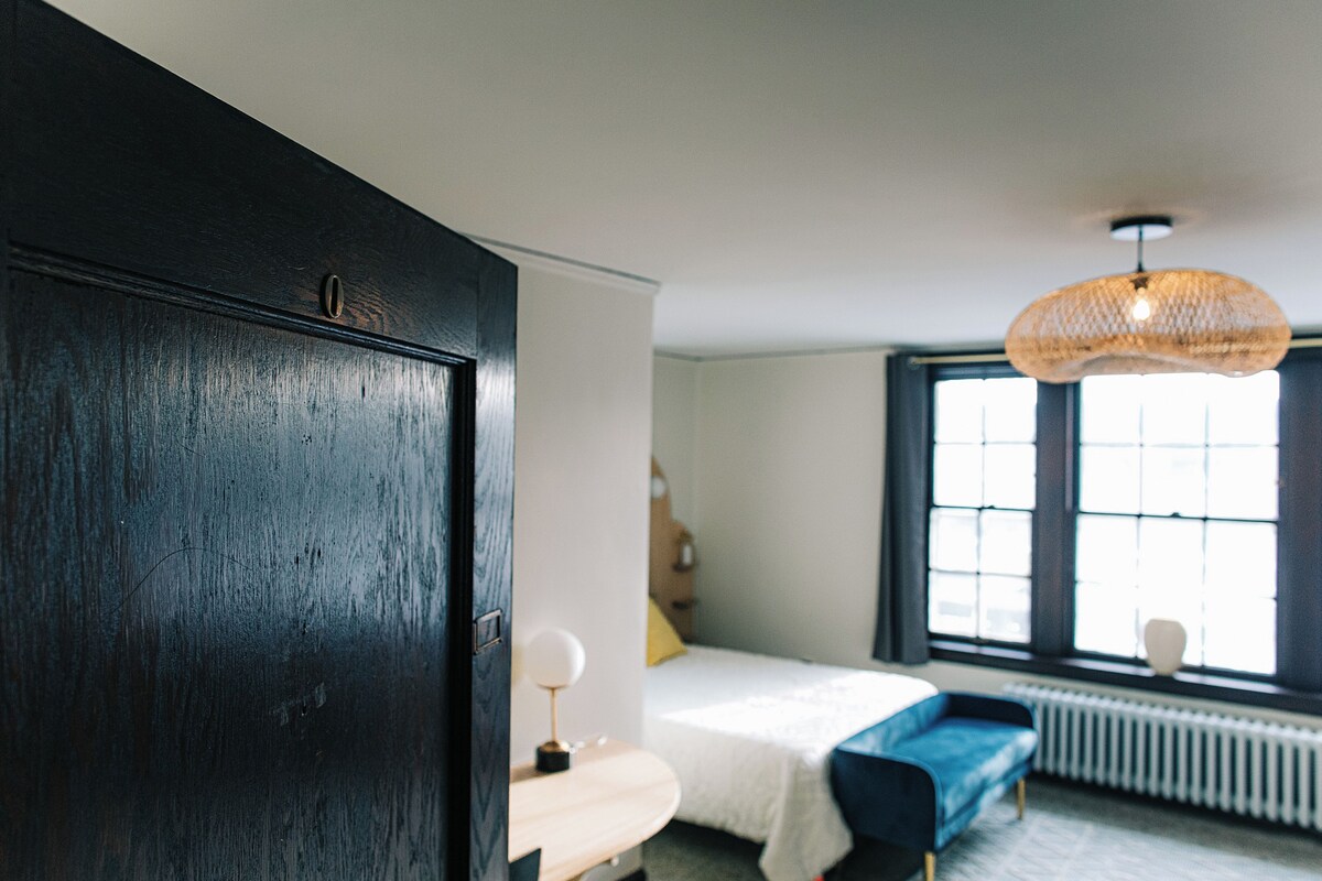 A bedroom with a bed, a bedside table, a lamp, a window with blinds, and a hanging light fixture.