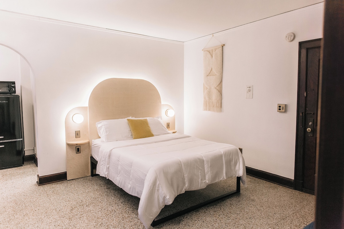 A bedroom with a bed, bedside lamps, a wall-mounted light, a door, and a wall-mounted switch.