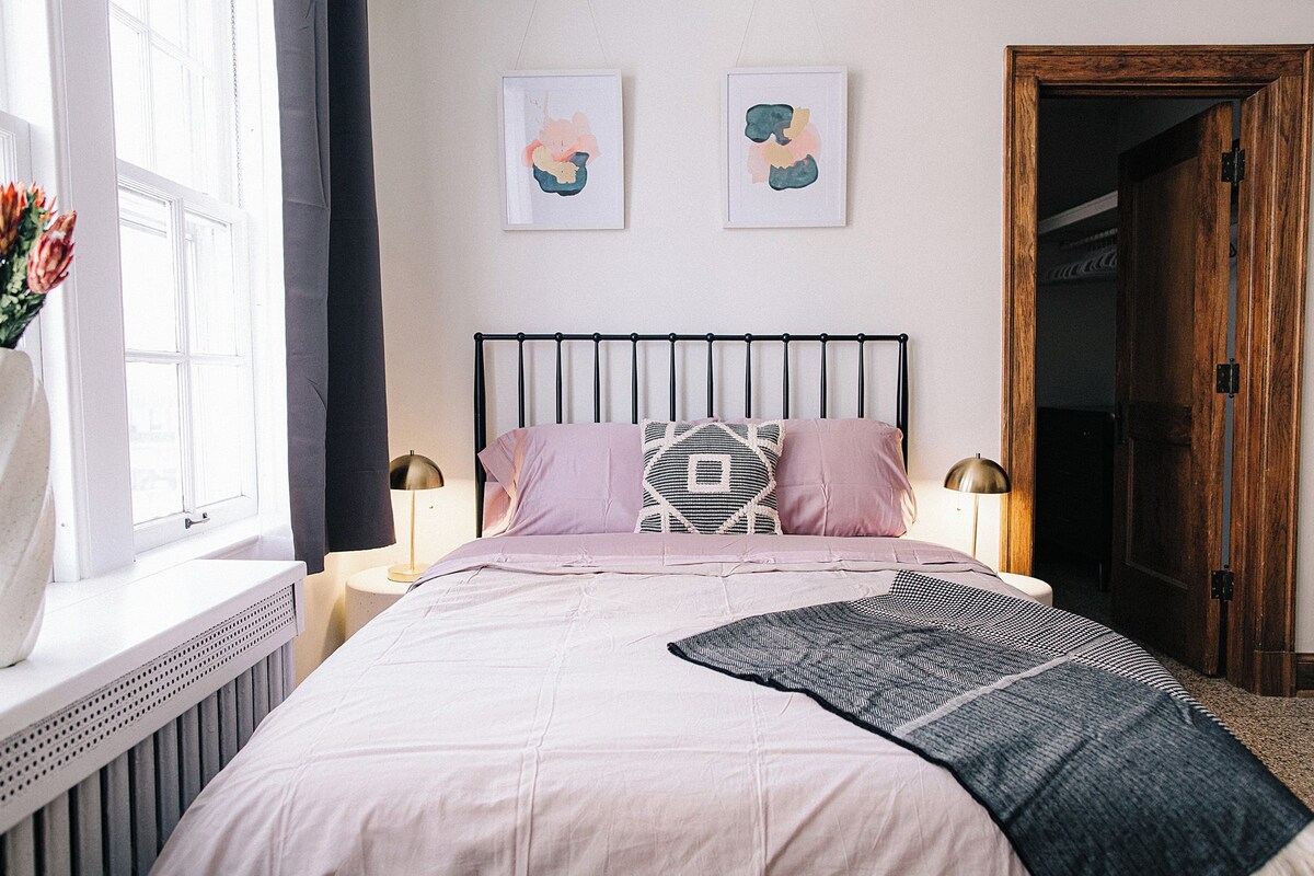 A bedroom with a bed, two bedside lamps, a radiator, a window, and two wall-mounted artworks.