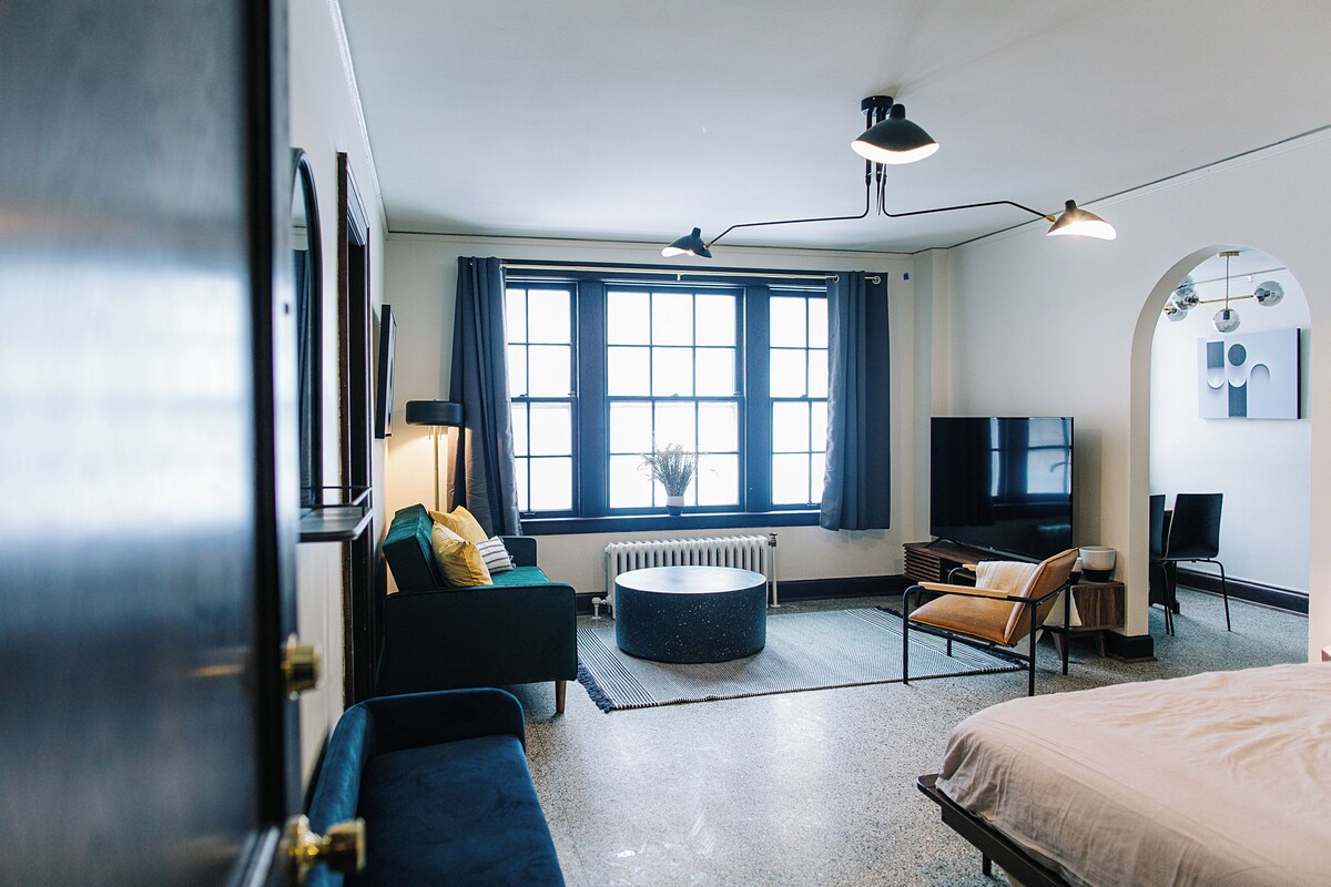 A hotel room with a bed, a sofa, a round ottoman, a television, and a window with curtains.