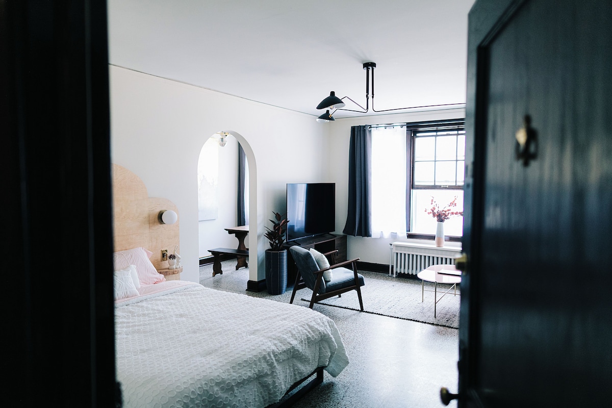A modern hotel room with a bed, a desk, a chair, a TV, and a window with curtains.