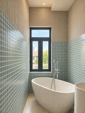 A bathroom with a freestanding bathtub, a window, and tiled walls. - The Mango Stay Thao Dien (An Khanh)