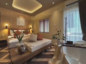 A hotel room with a bed, bedside tables, a sofa, a window with blinds, a potted plant, and a wall-mounted air conditioner. - The Mango Stay Thao Dien (An Khanh)