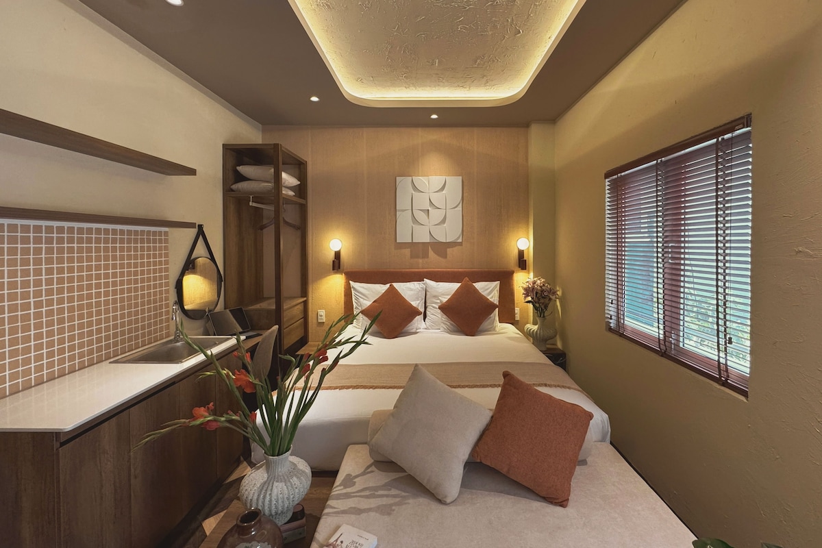 A modern hotel room with a large bed, bedside tables, a built-in wardrobe, a bathroom with a sink, and a window with blinds.