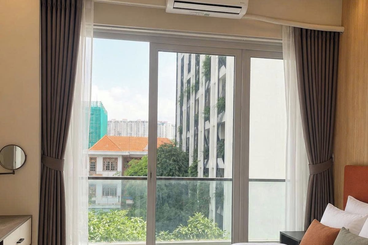 A hotel room with a bed, an air conditioning unit, a window with curtains, and a view of buildings outside.