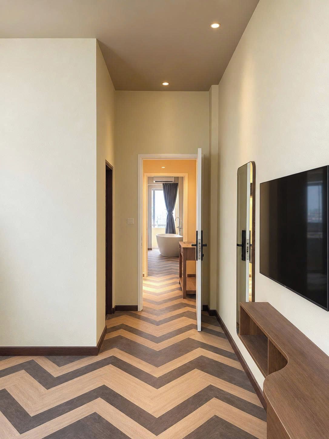 Un couloir d’hôtel avec un sol à motifs en chevrons, un téléviseur à écran plat fixé au mur et une vue sur une chambre dotée d’une baignoire et d’une fenêtre.