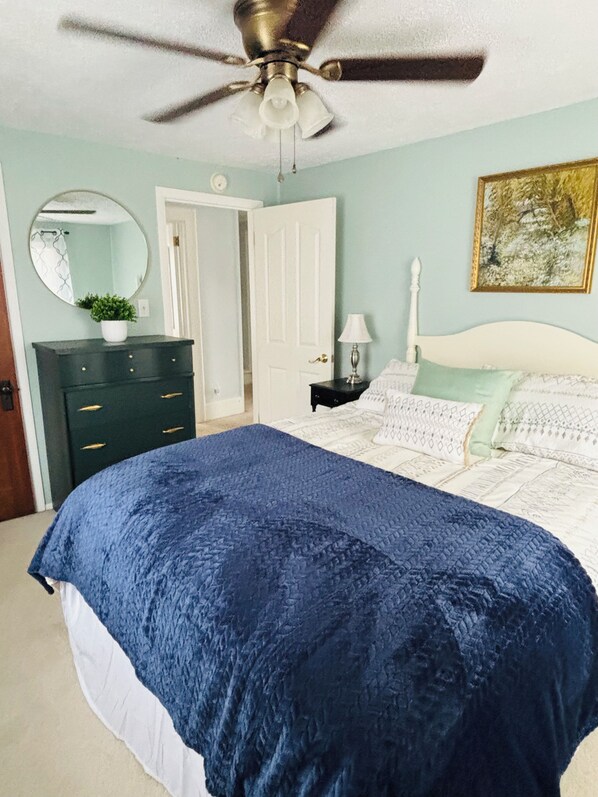 A bedroom with a bed, a ceiling fan, a dresser, a nightstand, a lamp, a mirror, and a door.