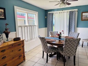 A dining area with a table set for four, patterned chairs, a wooden cabinet, and a window with a view of a building with a red roof. - It's a Wonderful House! (Indiana)