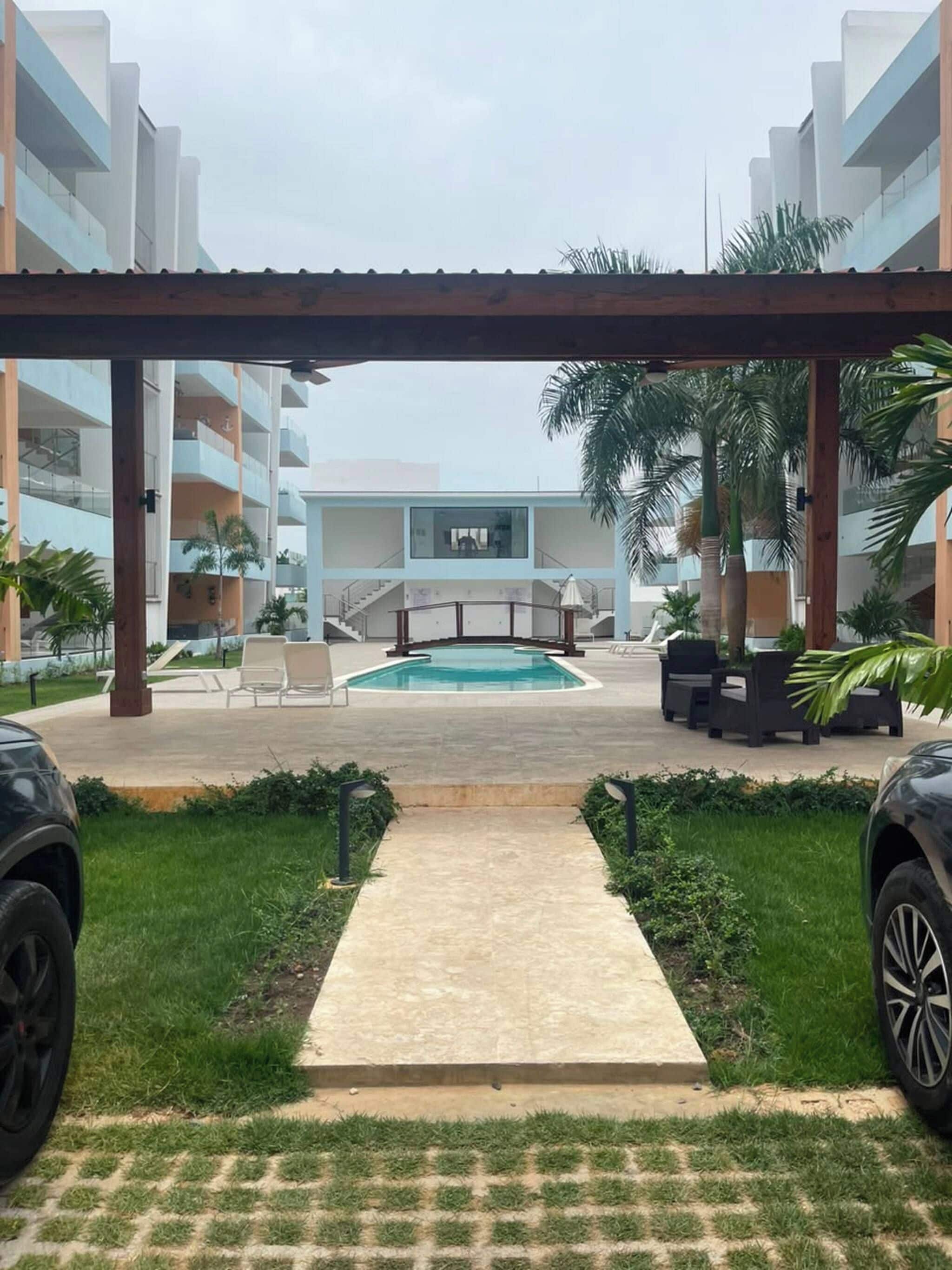 Zona de piscina con una pérgola, sillas de descanso y vista a un edificio moderno.