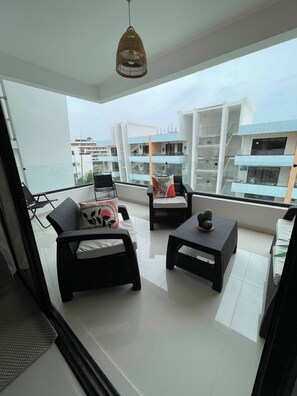 A balcony with wicker furniture, a hanging light fixture, and a view of modern buildings. - Bayahibe Sunshine 2BR Pool Close to the Beach (San Rafael del Yuma)