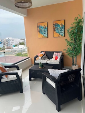 A balcony with wicker furniture, a potted plant, and artwork on the wall. - Bayahibe Sunshine 2BR Pool Close to the Beach (San Rafael del Yuma)