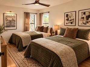 Une chambre d’hôtel avec deux lits, un ventilateur de plafond, une fenêtre avec des rideaux et des œuvres d’art accrochées aux murs.