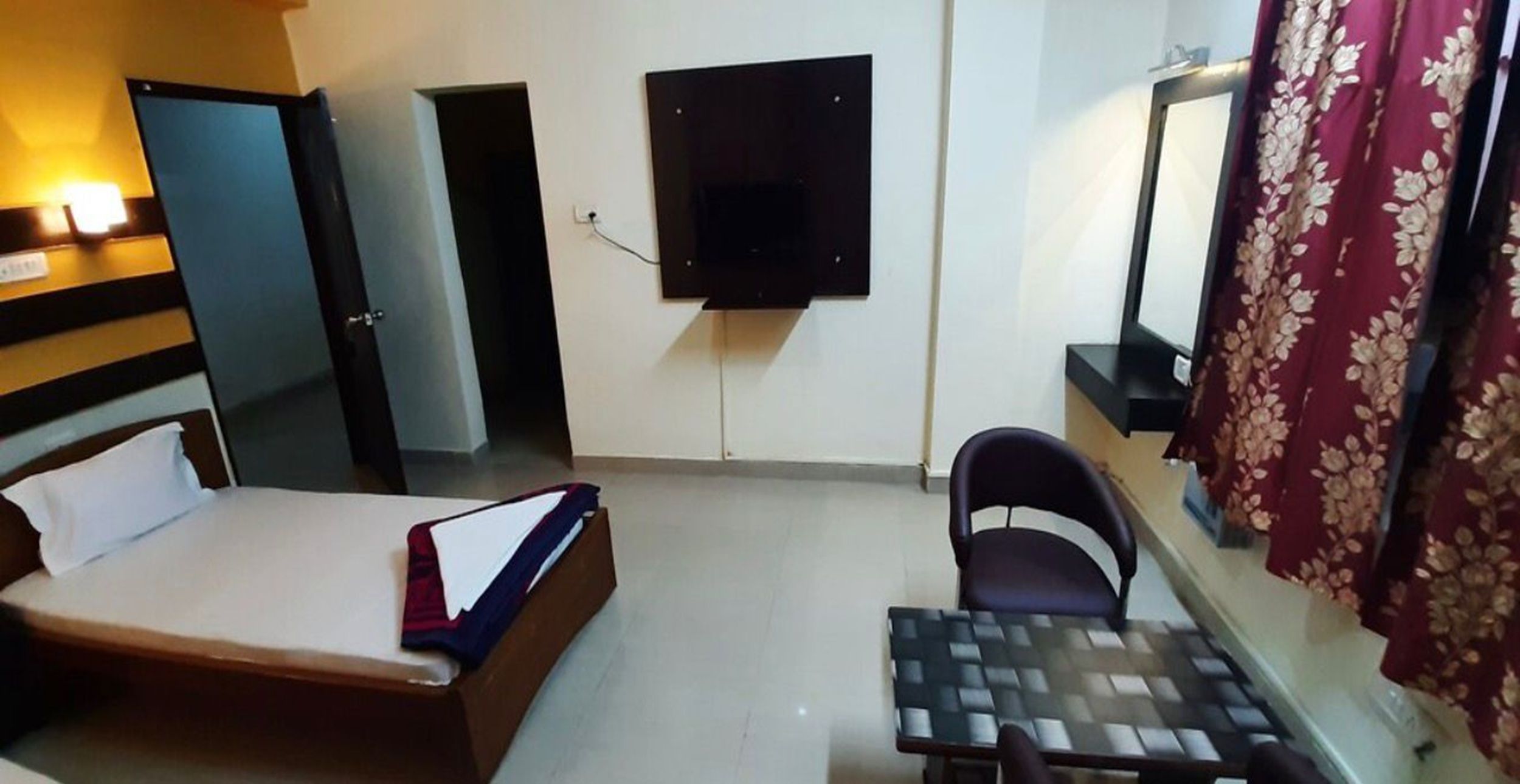 A hotel room with a bed, a desk, a chair, a TV mounted on the wall, and a door.