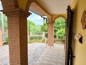 Área externa coberta com pilares de tijolo, piso de azulejos e vista para o verde.
