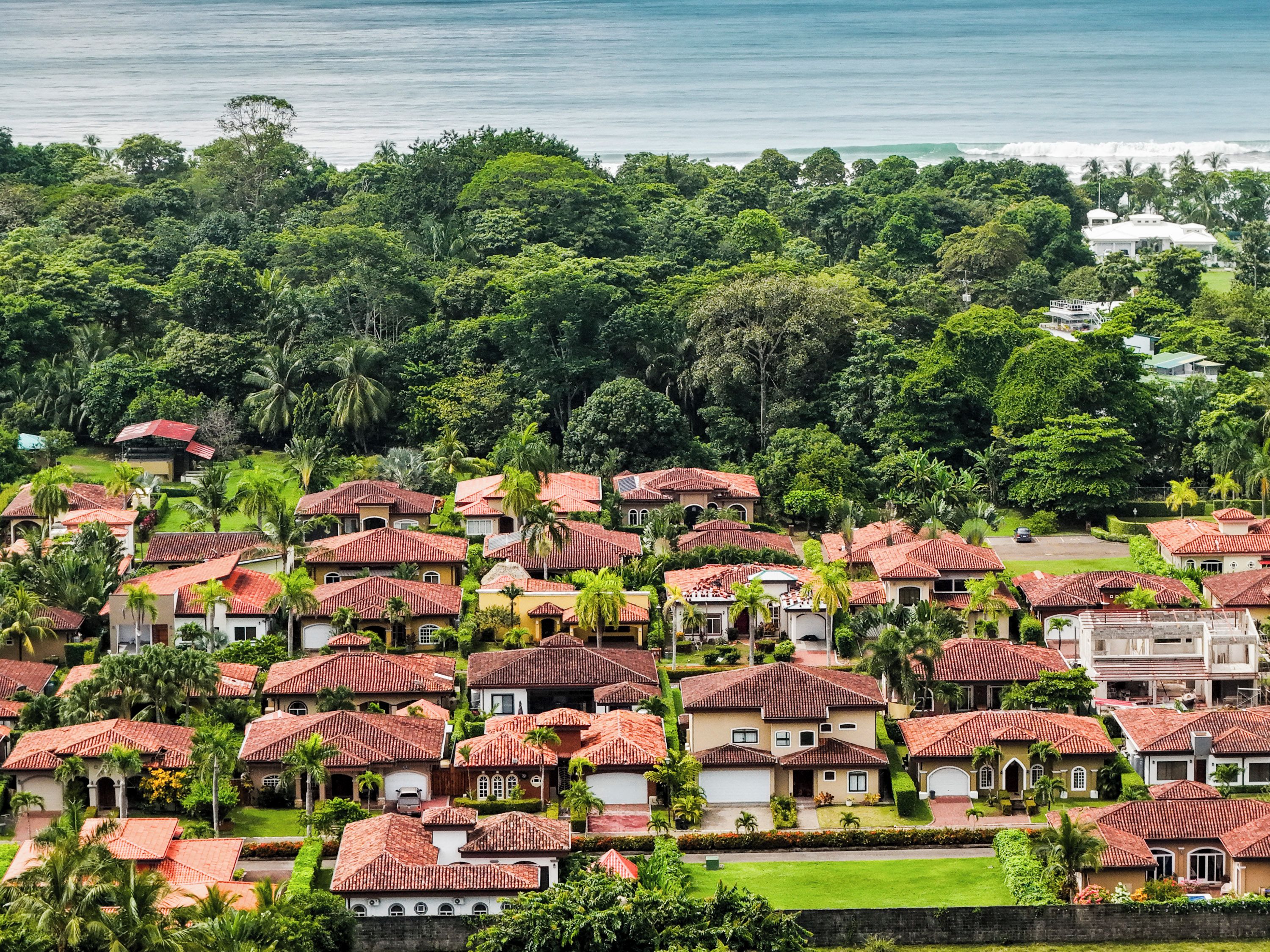 A coastal residential area with houses featuring red-tiled roofs, surrounded by lush greenery and a view of the ocean.
