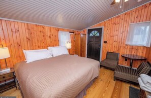 Ein Zimmer mit Holzwänden, einem Bett, einem Sessel, einem Tisch und einer Tür mit Fenster.