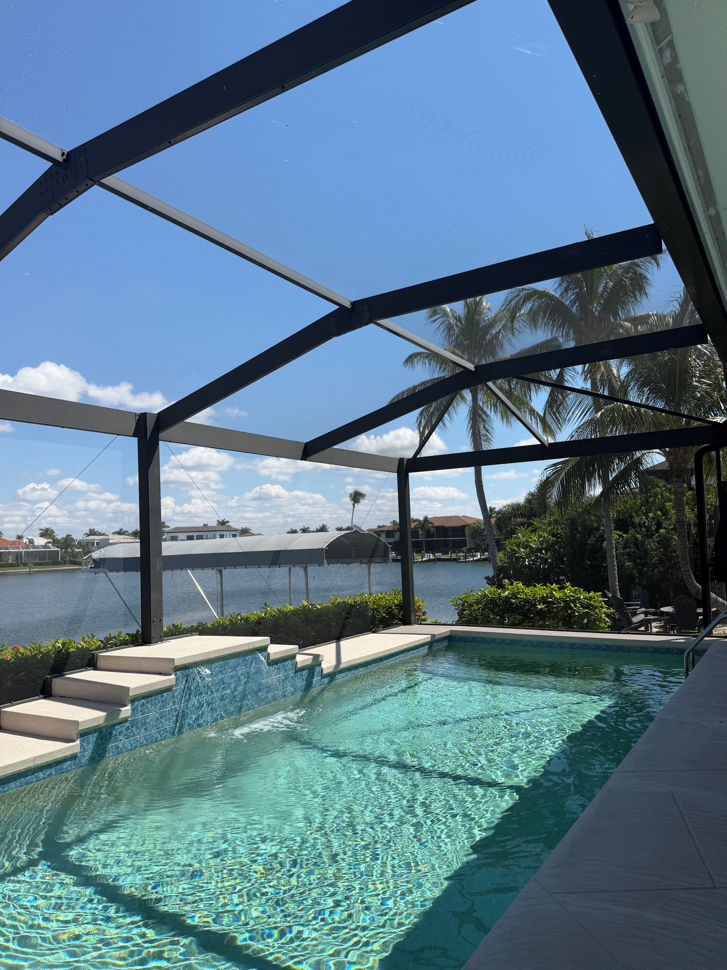 A swimming pool with clear water, surrounded by a covered area with metal beams, palm trees, and a view of a body of water.