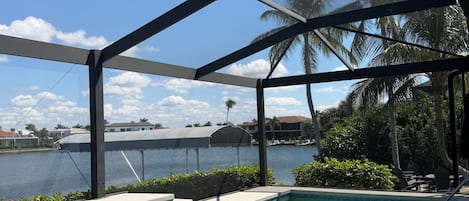 A swimming pool with clear water, surrounded by a covered area with metal beams, palm trees, and a view of a body of water.