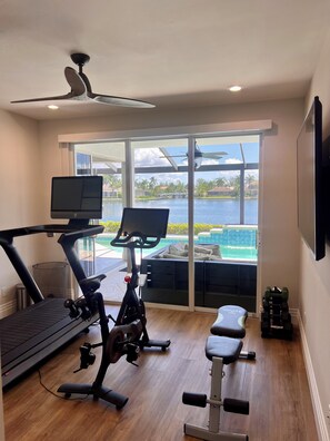 Ein Heimfitnessraum mit Laufband, Ergometer und Gewichtheckbank, mit Blick auf einen Pool und einen wolkenlosen Himmel.