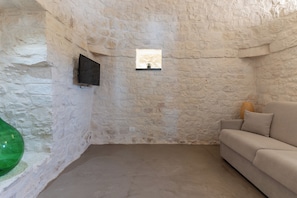 A room with a stone wall, a mounted flat-screen TV, a small window, and a beige sofa. - Luxury Trulli Zaccano Estate with Private Pool, Cisternino (Puglia)