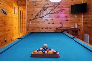 Una mesa de billar con un montón triangular de bolas de billar en una sala de madera con un televisor montado en la pared.
