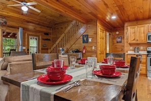 Interior de una cabaña de madera con una mesa de comedor dispuesta para cuatro personas, una escalera y una zona de cocina.