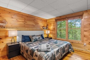 A wooden cabin bedroom with a bed, nightstands, and a large window. - Hot Tub • Game Room • 3 King Beds • Pet Friendly (Ellijay)