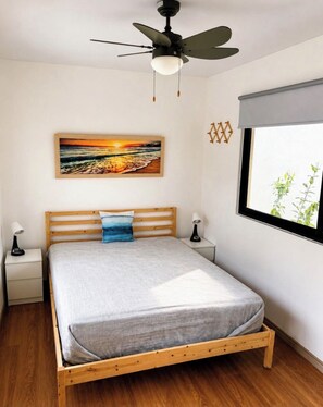 A bedroom with a wooden bed, a ceiling fan, a nightstand with a lamp, a window with a view of greenery, and a painting of a sunset. - CasaMara #2, tropical bungalow in Private Residential area walkable to the beach (Las Terrenas)